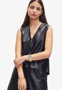 Top sans manches en cuir noir avec un décolleté en V et une coupe ample. Présente une texture lisse et un design en couches. Le modèle porte une montre en or et des bijoux.