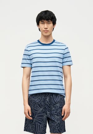 Młody mężczyzna ubrany w jasnoniebieską, paskowaną koszulę z krótkim rękawem oraz granatowe szorty z białym wzorem liści, stojący na tle gładkiej ściany.