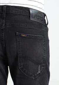 Černé džínové kalhoty z denimu s výraznou logo nášivkou na zadním pásku a nenápadnou značkovou šťítkem na zadní kapse.