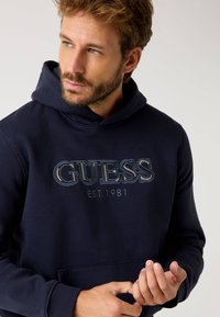 Sötétkék kapucnis pulóver elülső kenguruzsebbel, a mellkason texturált hímzéssel ellátva, melyen a "GUESS EST. 1981" felirat található. Puha anyagú.