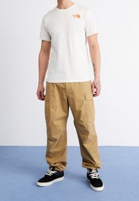 Béžové tričko s malým oranžovým logem, doplněné khaki cargo kalhotami s bočními kapsami. Černé tenisky s bílou podrážkou.