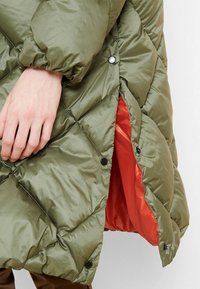 Cappotto trapuntato verde oliva con una finitura lucida, dotato di bottoni a pressione neri e di una fodera arancione a contrasto.