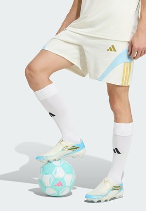 Persona che indossa scarpe da calcio bianche e azzurro chiaro, calzini bianchi e pantaloncini bianchi con accenti dorati e blu, controllando un pallone da calcio azzurro chiaro e rosa.