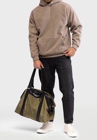 Olivengrøn duffelbag med sorte stropper, lynlås og "rebel" logo. Båret af en person i en beige sweatshirt og mørke jeans. Hvide sneakers.