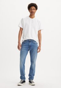 T-shirt en coton blanc, jean en denim bleu, coupe décontractée, délavé clair, coutures contrastantes, porté avec des baskets noires à semelles blanches.