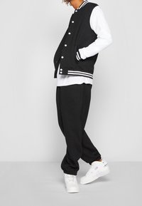 Giacca varsity nera con maniche bianche, polsini a righe e chiusura con bottoni, abbinata a pantaloni jogger neri e sneakers bianche. Aspetto semplice e sportivo.