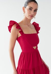 Vestido rojo con tirantes con volantes, escote cuadrado y una abertura en forma de gota. La tela parece ligera y texturizada. Diseño de falda en capas.