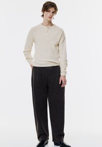 Cămașă lungă cu mâneci ribbed de culoare crem, cu un guler henley, asociată cu pantaloni wide-leg de culoare gri închis, completată cu botoși negri până la gleznă și un lanț metalic.