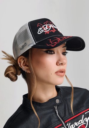 Młoda kobieta z wieloma kolczykami w uszach i pierścieniem w nosie, nosząca czarno-białą czapkę Ed Hardy oraz czarną kurtkę z czerwono-białymi napisami.