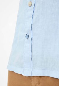 Camicia di lino azzurro chiaro con dettagli in bottoni, cuciture visibili e orlo dritto. Il tessuto ha un aspetto morbido e textured.