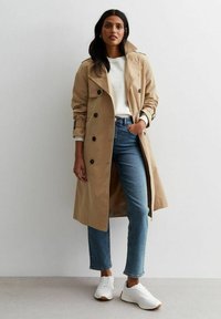 Beige trenchcoat med dubbelknäppning, matchad med en vit topp och blå jeans. Vita sneakers fulländar looken.
