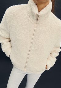 Kremna flis jakna z visokim ovratnikom, celim zadrgo na sprednji strani in teksturirano, puhasto površino. Dolgi rokavi z elastičnimi manžetami.