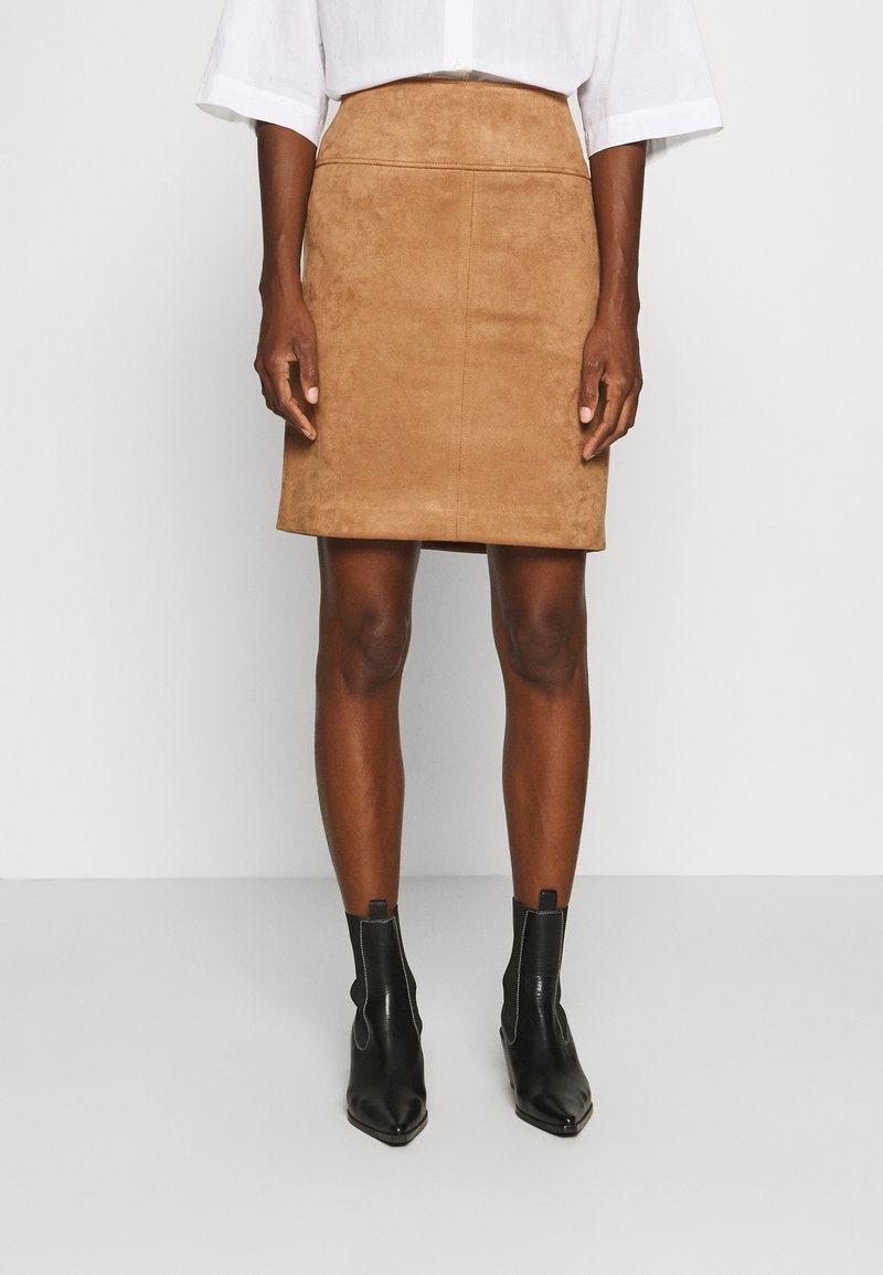 Jupe en daim couleur tan, longueur genou, avec un design ajusté et des détails de couture verticales, associée à des bottines noires à bout pointu.