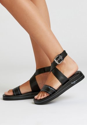 Sandalias de cuero negro con tiras cruzadas, una correa en el tobillo con hebilla, una suela texturizada en negro y líneas de acento blancas en el borde.