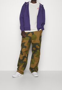 Sudadera morada con cremallera, camiseta blanca con logo rojo, pantalones de carga camuflados en verde y marrón, zapatillas blancas. Diseño simple y funcional.