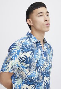 Camicia a maniche corte in tessuto leggero, caratterizzata da una stampa tropicale blu e verde con ananas e foglie di palma su uno sfondo crema.