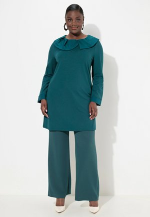Abito tunica color teal con un ampio colletto a pieghe, maniche lunghe e pantaloni a gamba larga coordinati. Tessuto liscio, abbinato a tacchi di colore chiaro.