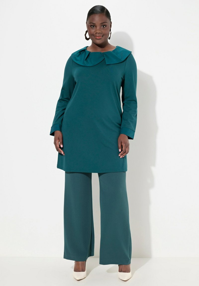 Abito tunica color teal con un ampio colletto a pieghe, maniche lunghe e pantaloni a gamba larga coordinati. Tessuto liscio, abbinato a tacchi di colore chiaro.