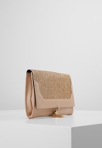 Beige laukku, jossa on kimaltava kultainen läppä, tekstuurinen pinta ja koristeellinen pallohäntä kiinnityksessä.