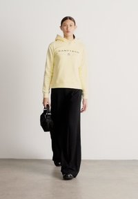 Žltá mikina s kapucňou a predným logom, v kombinácii s voľnými čiernymi nohavicami. Modelka drží malú čiernu kabelku a stojí na neutrálom pozadí.