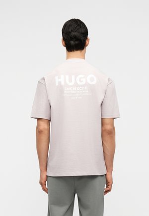 Mann med mørkt hår iført lys beige oversized T-skjorte med hvit "HUGO"-logo og tekst på baksiden, kombinert med grå bukser, står foran hvit bakgrunn.