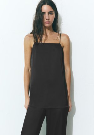 Jeune femme aux longs cheveux foncés portant un haut noir ample sans manches et un pantalon noir, debout contre un mur blanc.