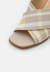 Beżowe plecione sandały wsuwane z żółtymi i białymi akcentami. Posiadają płaską podeszwę, krzyżowy wzór i górę o gładkiej, teksturowanej powierzchni.