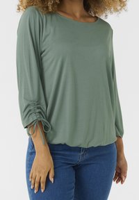 Zelený, dlouhý rukáv top vyrobený z měkkého materiálu, s výstřihem ve tvaru lodičky a skládacími rukávy se šňůrkami. V kombinaci s modrými džínovými kalhotami.