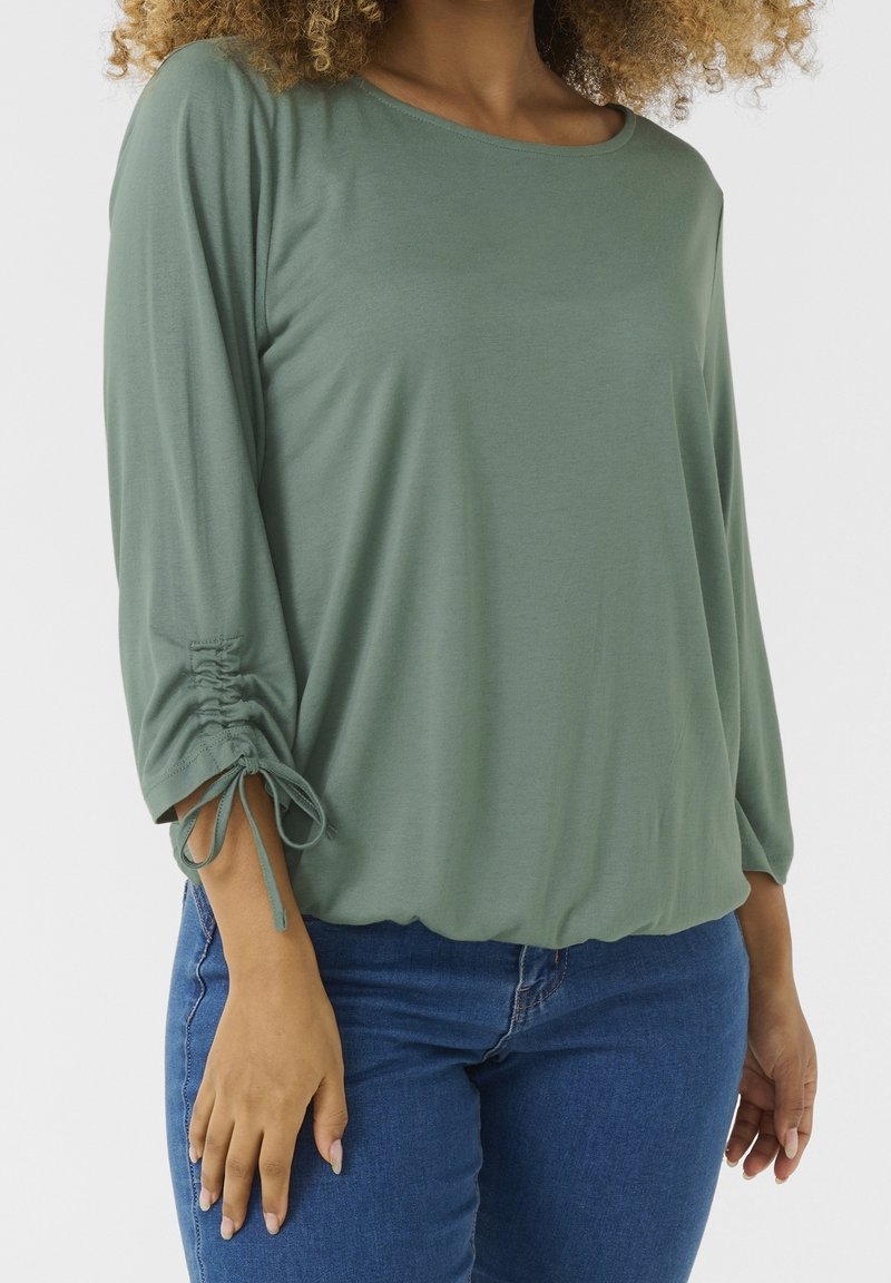 Zelený, dlouhý rukáv top vyrobený z měkkého materiálu, s výstřihem ve tvaru lodičky a skládacími rukávy se šňůrkami. V kombinaci s modrými džínovými kalhotami.