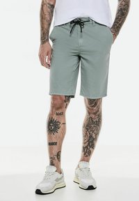 Shorts gris clair avec un taille élastique à cordon, poches avant et texture lisse ; associés à des baskets blanches et des tatouages visibles.