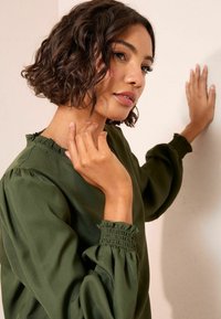 Zöld, hosszú ujjú blúz sima anyagból, húzott mandzsettával és fodros nyakkivágással, semleges háttér előtt bemutatva.