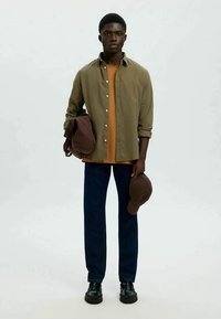 Young man stands facing forward, wearing olive shirt over orange tee, dark pants, black shoes, holding brown hat and jacket against white background.