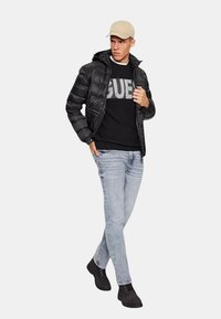 Giacca puffer nera con cappuccio, felpa nera con grafiche, jeans azzurri chiari e stivali neri. Cappello beige chiaro sulla testa.