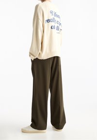 Beige neulottu neule, jossa sininen painettu teksti selässä, yhdistetty tummanvihreisiin leveälahkeisiin housuihin ja kermaan sävyisiin lenkkareihin.