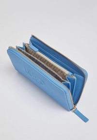 Portefeuille zippé bleu clair, ouvert pour montrer plusieurs compartiments, emplacements pour cartes, et un intérieur beige avec une doublure en tissu de marque.