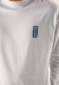 Langermet hvit t-skjorte i bomull med en blå rektangulær logo med teksten "MT FUJI" og et blomsterdesign. Glatt tekstur, enkelt design.
