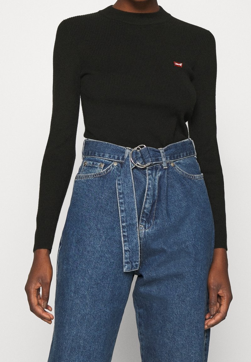 Personne portant un top noir côtelé à manches longues avec un petit logo rouge et un jean bleu taille haute avec une ceinture en denim assortie.