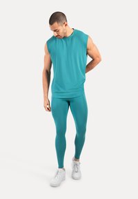 Mężczyzna w turkusowej koszulce bez rękawów i pasujących legginsach, w białych sneakersach, patrzy w dół z jedną ręką za plecami.