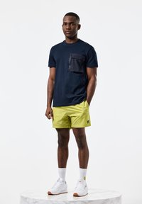 Tricou bleumarin cu buzunar cu fermoar, asortat cu pantaloni scurți galbeni strălucitori. Modelul poartă adidași albi și șosete albe pe o bază din marmură.