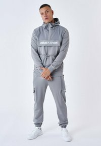 Tuta grigia realizzata in materiale morbido, con giacca con cappuccio dotata di tasca frontale e cordino regolabile, accompagnata da joggers aderenti abbinati.