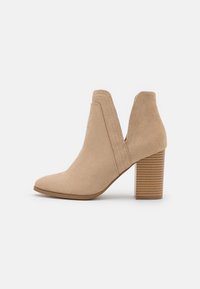 Beige Wildleder-Ankle-Boot mit spitzem Vorderteil, V-förmigem Ausschnitt und einem gestuften Absatz mit horizontalen Streifen. Glatte Textur und klare Linien.