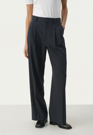Navyblau gestreifte Hose mit hoher Taille, weitem Bein und tailliertem Design. Aus einem glatten Stoff gefertigt. Getragen mit einem weißen T-Shirt und schwarzen Schuhen.