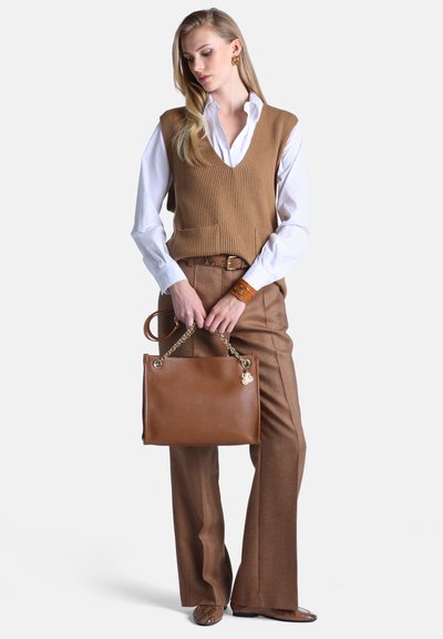 Femme portant une chemise blanche et un gilet en maille marron avec un pantalon marron assorti, tenant un sac à main en cuir marron avec des anses en chaîne dorée.