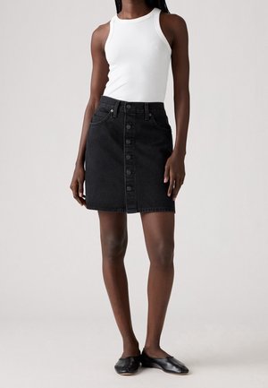 Femme portant un haut blanc sans manches, une jupe en jean noire avec boutons sur le devant et des chaussures plates noires, debout devant un fond uni.