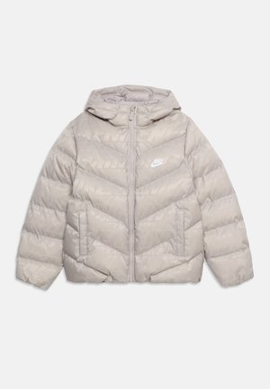 Vaaleabeige Nike toppatakki hupulla, edessä vetoketju, kaksi sivutaskua ja valkoinen logo rinnassa, aseteltu tasaisesti valkoiselle taustalle.