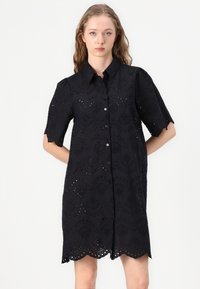 Abito nero a camicia con maniche corte a scallop e chiusura frontale con bottoni. Presenta intricati motivi floreali a intaglio. Tessuto leggero, lunghezza al ginocchio.