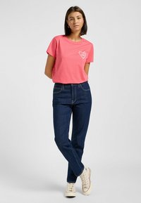 T-shirt en coton rose clair avec un motif de cœur, associé à un jean en denim bleu foncé. Modèle portant des baskets beiges. Design et coupe simples.