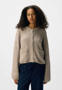 Beige gestrickte Strickjacke aus weichem Material, mit langen Ärmeln, zwei Vordertaschen und goldenen Knopfverschluss.