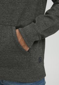 Mørkegrå sweatshirt med ribbede manchetter og kant, der har en kengurulomme. Lille mærke med teksten "DNMWRKRS" på den nederste side.