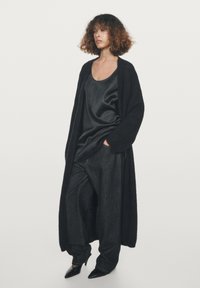 Lange zwarte cardigan van een zachte stof over een donker satijnen tanktop en losse donkere jeans, aangevuld met puntige zwarte hakken.